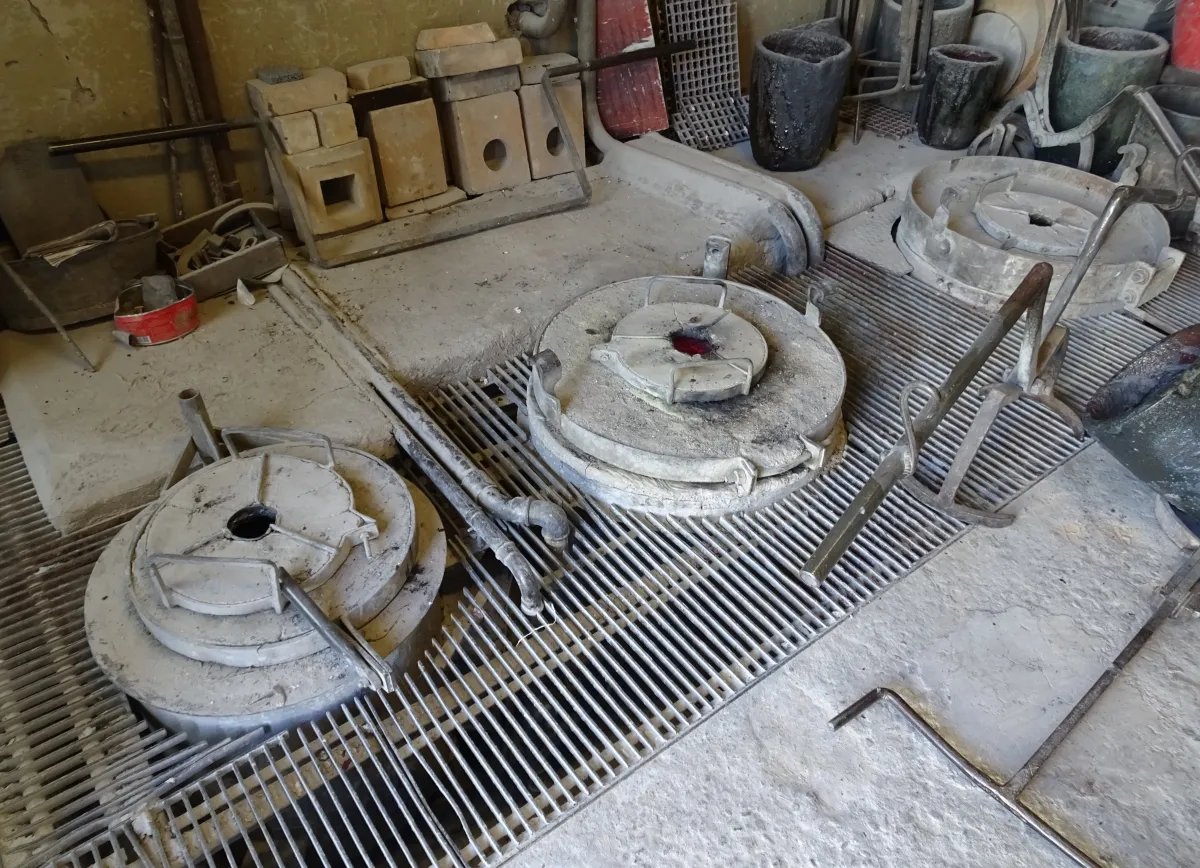 Runda, gråa formar med hål i mitten ligger på ett metallgaller i golvet. Längs väggen bakom står svarta och gröna deglar och staplade eldfasta stenar.