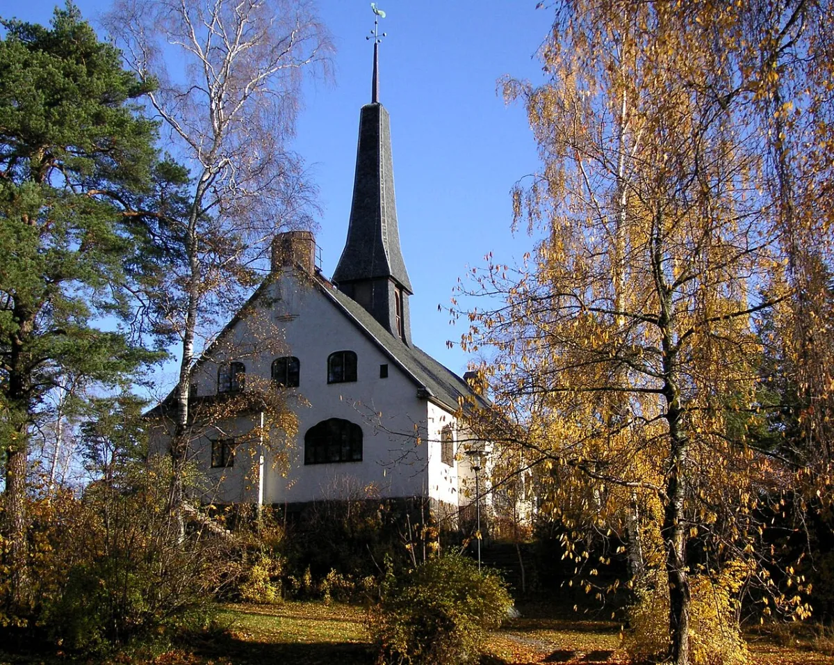 Enskede kyrka, vit med svart tak och klocktorn mot en blå himmel med träd runtomkring.