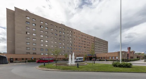 Ett långt brunt tegelhus i tio våningar med en lägre byggnad framför, där entrén ligger. I en rondell framför huset står en fontän, och vid entrén står en röd buss och en vit minibuss.