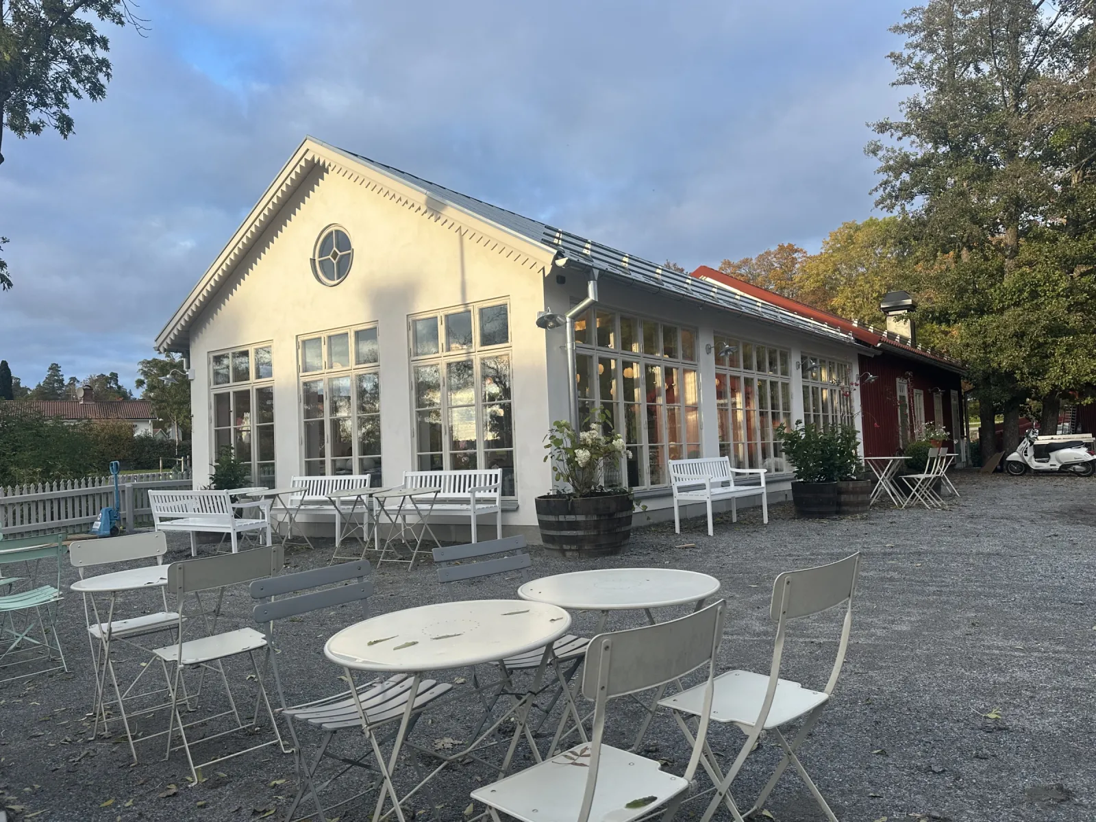 En nybyggd, låg, vit byggnad med stora spröjsade fönster och ett runt fönster i gaveln, Enskedeparkens bageris nya orangeri. Framför står vita kaféstolar och bord.