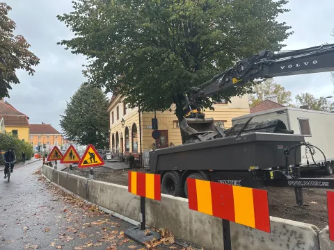 En grävmaskin på ett släp bakom en rad betongblock och vägarbetsskyltar. Bakom den står ett stort lövträd och ett gult hus med rundbågade fönster. Till vänster cyklar någon på gatan.