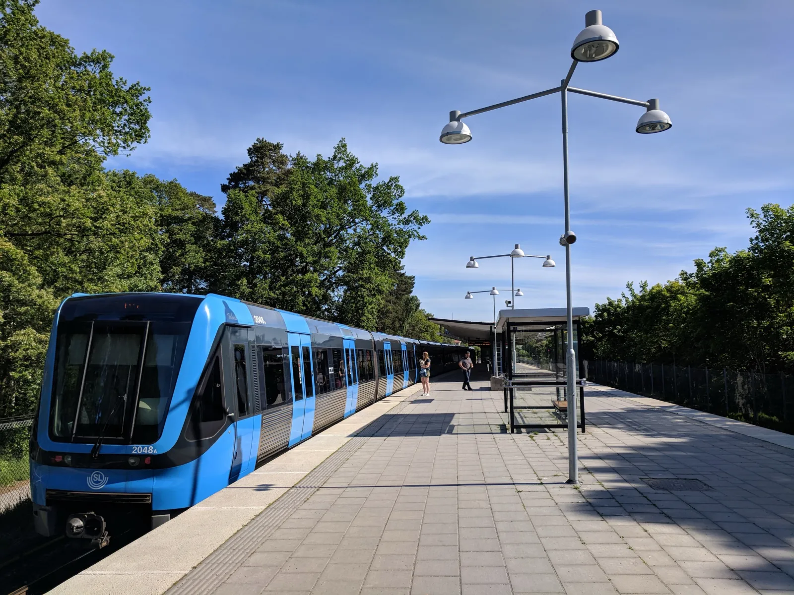 Ett blått tunnelbanetåg står vid en perrong ovan jord, omgiven av höga lövträd. På perrongen står ett väntskjul och lyktstolpar med tre lampor, och längre bort väntar två personer.