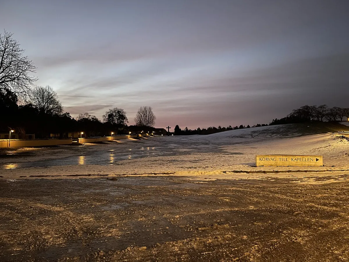Skogskyrkogården i gryningen en vintermorgon, med frost och snö på marken och lyktor längs vägen. Långt bort syns granitkorset mot en rosa himmel, och en skylt visar körvägen till kapel