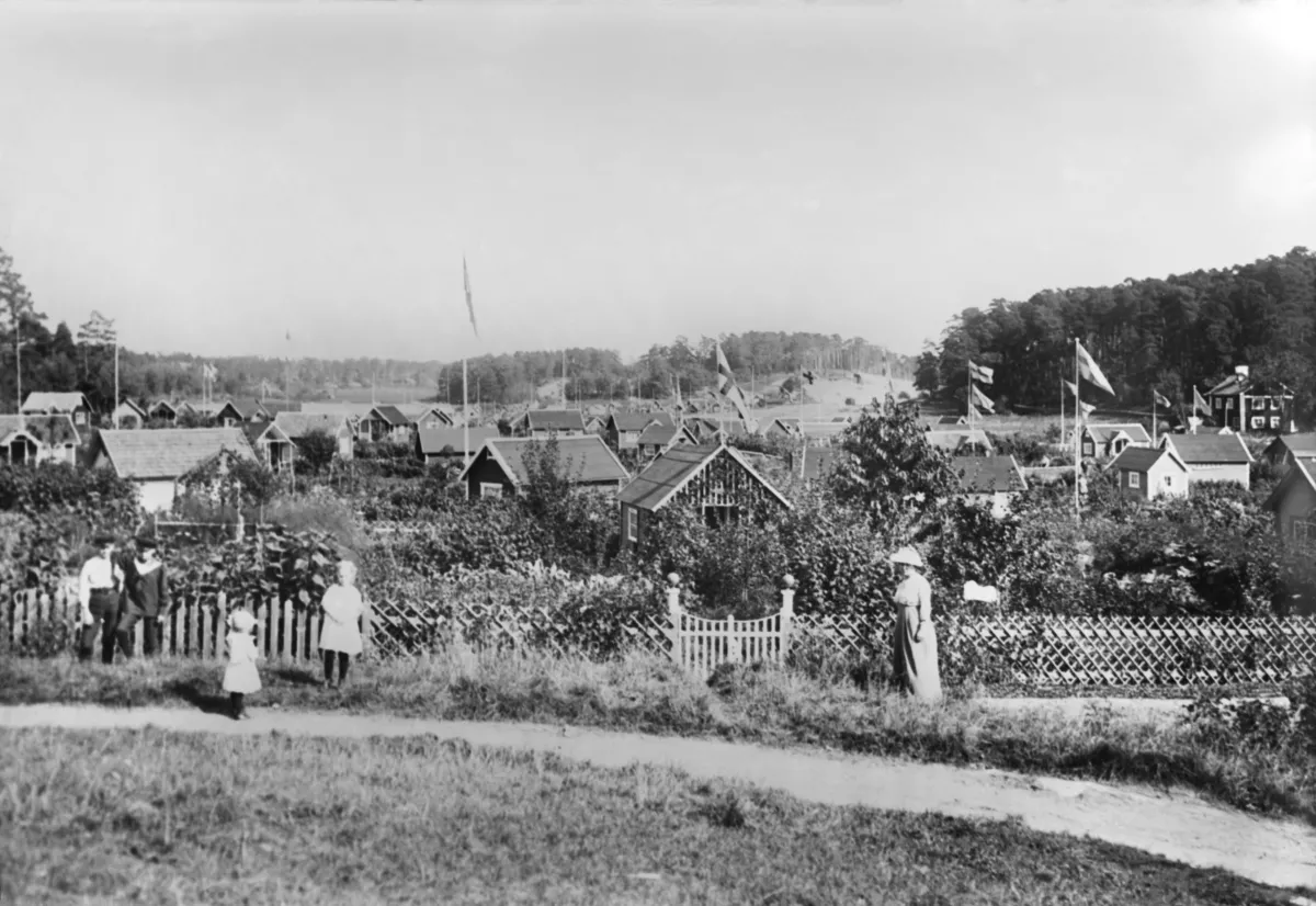 Svartvitt foto av små kolonistugor med flaggstänger bakom ett spjälstaket. På en grusgång står en kvinna i lång klänning och hatt, och vid staketet några män och två barn. Bakom stugorna reser sig skogsklädda höjder.