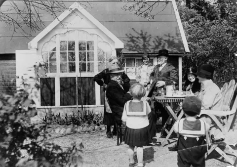 Svartvitt foto av en familj vid ett kaffebord framför en kolonistuga med ett stort spröjsat fönster. Två barn i sjömanskläder står i förgrunden.