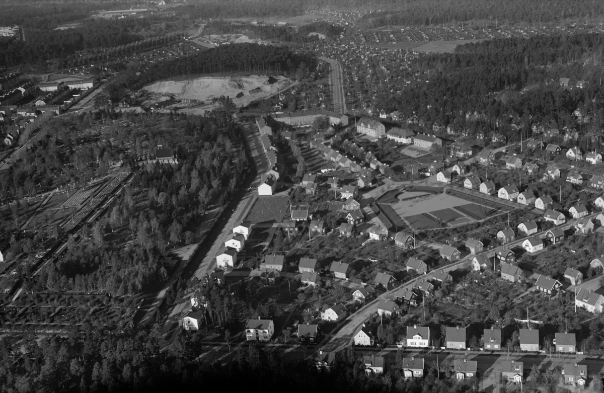 Svartvitt flygfoto över villor och trädgårdar. En väg går rakt upp genom bilden mot ett stort ljust grustag i en skogsklädd ås. Till höger ligger tätt med små hus och trädgårdar.