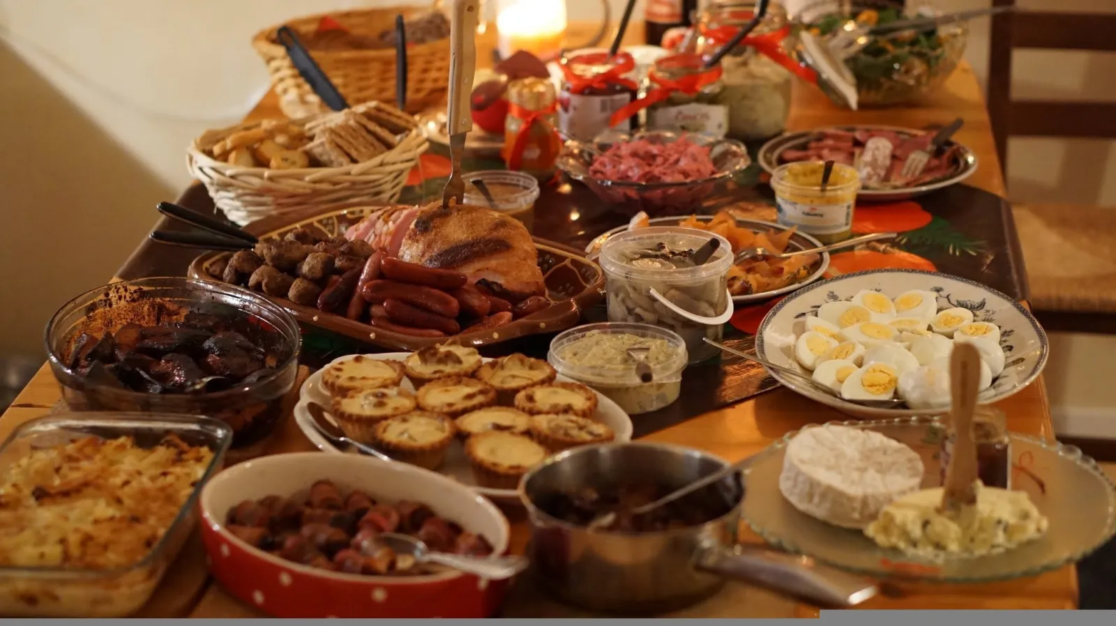 Ett julbord på ett träbord med Janssons frestelse, köttbullar, prinskorv, små pajer, ägghalvor, rödbetssallad och ost. Längst bak står en brödkorg och ett tänt ljus.