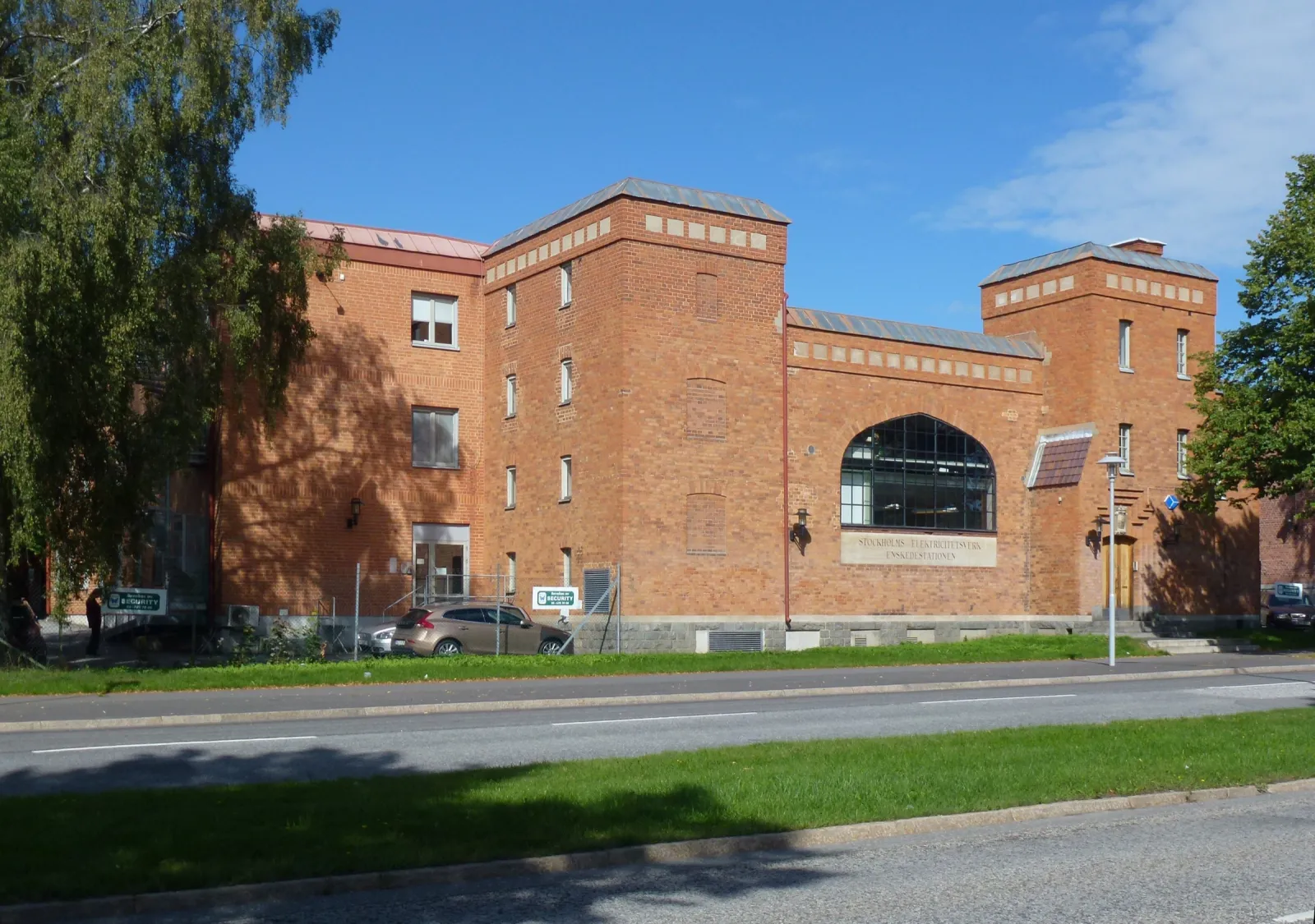 Ett tegelhus i rött med ett stort välvt fönster och en ljus skylt under det, sett från andra sidan Enskedevägen. Till vänster en högre del av huset och en björk, i förgrunden gräsremsor och körbanor.