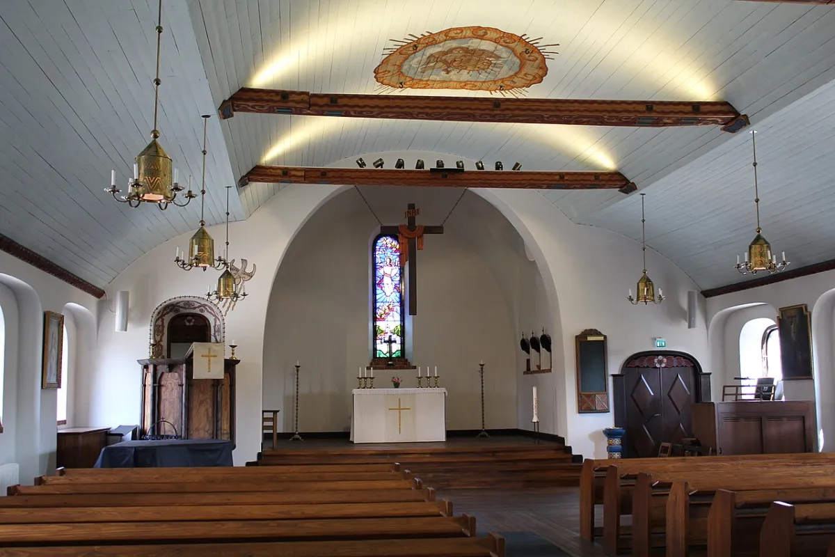 Kyrkorummet i Enskede kyrka sett mot koret. Rader av träbänkar leder fram till altaret, lampor i mässing hänger från det vita trätaket, och bakom altaret sitter ett färgat glasfönster och et
