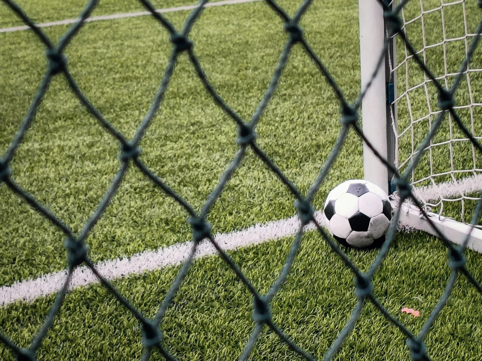 En svartvit fotboll ligger vid stolpen i ett mål på en konstgräsplan, fotograferad genom ett nät.