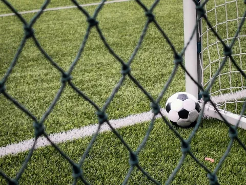 En svartvit fotboll ligger vid stolpen i ett mål på en konstgräsplan, fotograferad genom ett nät.