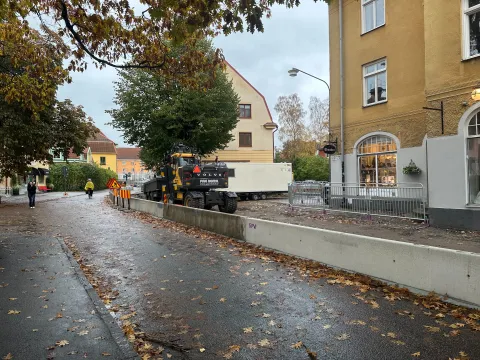 En gata med höstlöv, där en rad betongblock skiljer gatan från en uppgrävd yta med en grävmaskin och en vit bod.