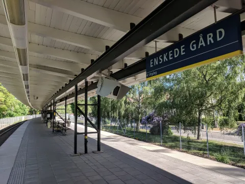 En tom perrong under ett vitt trätak med en blå skylt där det står Enskede gård. Bakom ett nätstängsel syns björkar, en blommande buske och en gata.