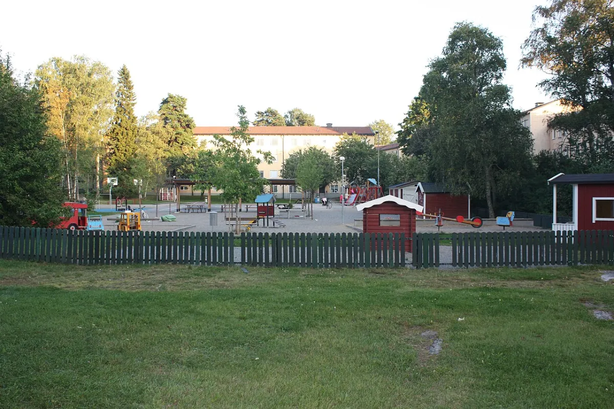 Parkleken Odlarängen i Tallkrogen bakom ett grönt staket, med små röda lekstugor och lekredskap på en grusplan mellan lövträd.