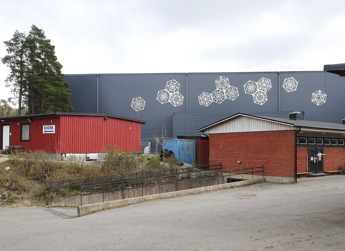 Gubbängens bandyhall, en stor grå hall med vita snöflingemönster på fasaden. Framför står en röd träbod med skylten Kiosk och en låg byggnad i rött tegel.
