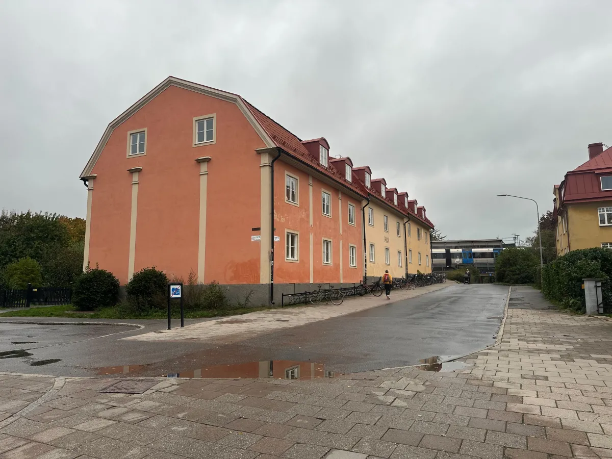 Ett laxrosa trevåningshus med brutet tak och takkupor längs en våt gata. I bakgrunden syns ett tunnelbanetåg.