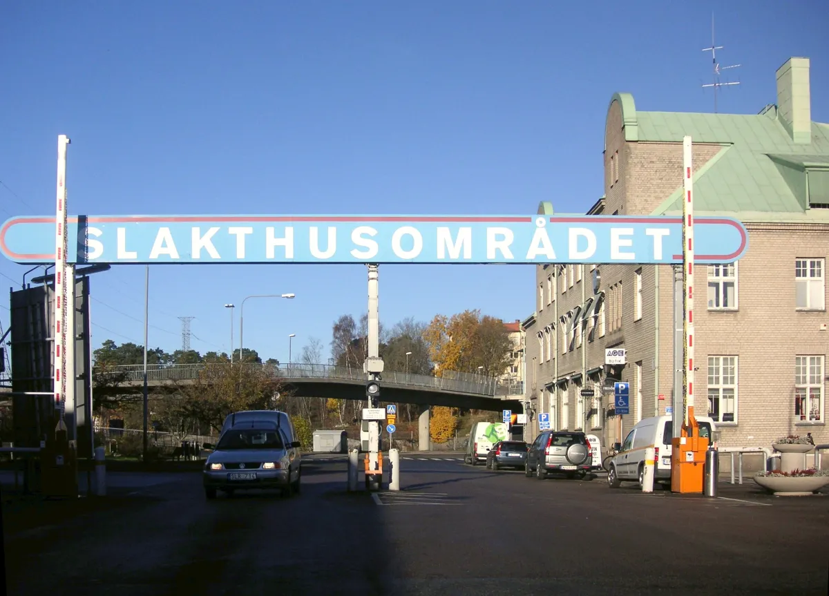 Infart med bommar under en stor skylt med texten Slakthusområdet. Till höger ett tegelhus med grönt tak