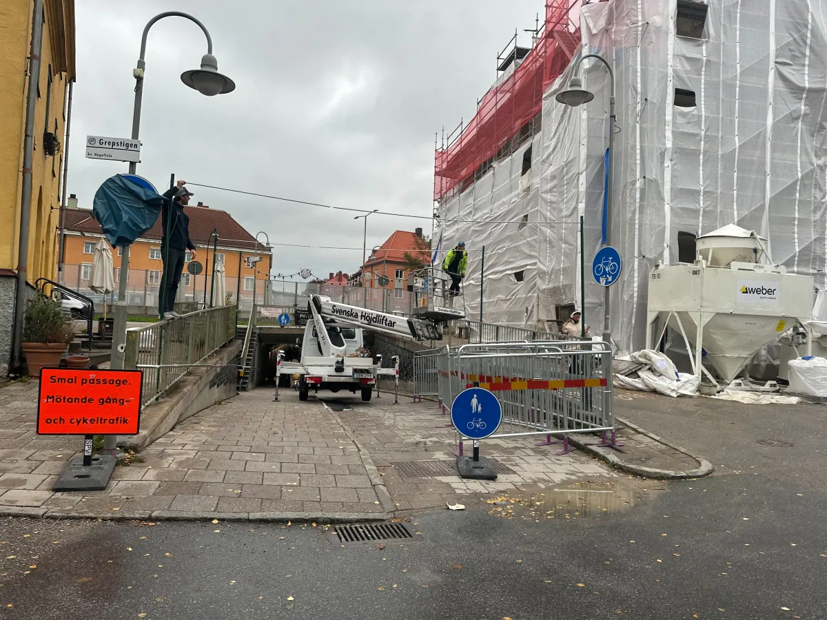 En gångväg som sluttar ner mot en tunnel, med en vit liftbil i gången och en arbetare i varselkläder uppe i liften. Till vänster står en orange skylt om smal passage, och till höger ett avspärrat hus inklätt i byggplast.