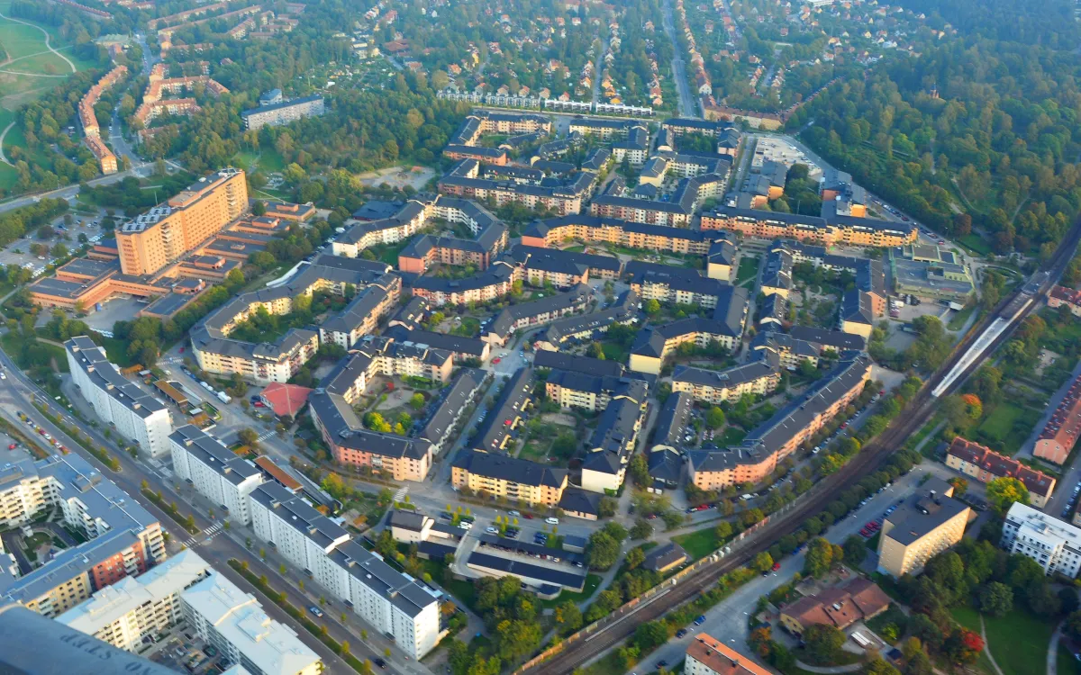 Flygfoto över ett bostadsområde med gula och bruna hus kring gröna gårdar. Till vänster står ett högt brunt skivhus med låga byggnader omkring, och till höger går tunnelbanan.