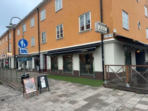 Ett orange hörnhus med en pizzeria i bottenvåningen och en trädäcksservering. Till vänster står blå vägmärken och skyltar om ersättningsbussar.
