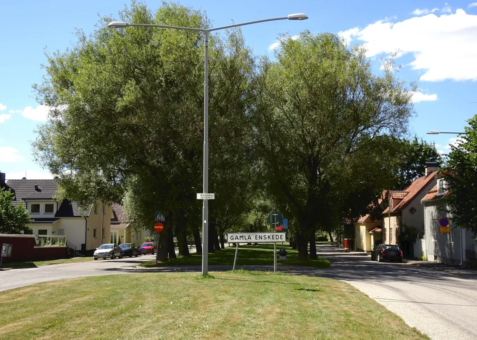 En gräsplätt med en skylt där det står Gamla Enskede, en gatlykta och höga lövträd. Gatan delar sig runt gräset, och på båda sidor står villor med tegeltak och parkerade bilar.