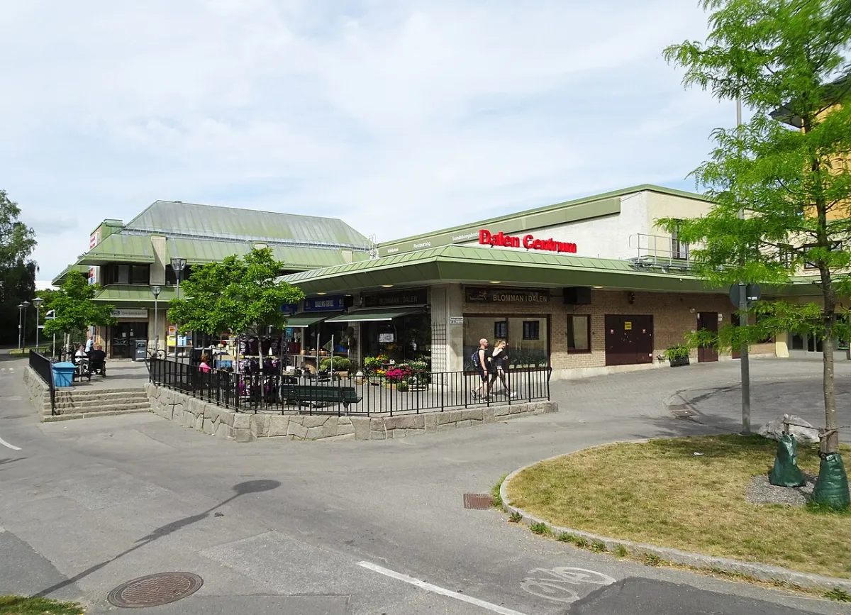 Dalens centrum en sommardag, med låga byggnader med gröna tak och skylten Dalen Centrum. Människor går på trottoaren framför, och till höger står ett träd.