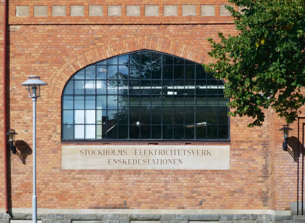 Ett stort välvt fönster med svarta spröjs i en tegelfasad. Under fönstret en ljus skylt med texten Stockholms Elektricitetsverk Enskedestationen. Till vänster en gatlykta och till höger grenarna från ett träd.