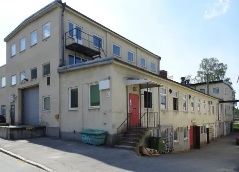 Ett ljust putsat verkstadshus i två och tre våningar med en hög port och en röd dörr högst upp på en trappa. Vid dörren sitter skyltar med 44 och C, och till höger fortsätter en lång, låg länga längs gatan.