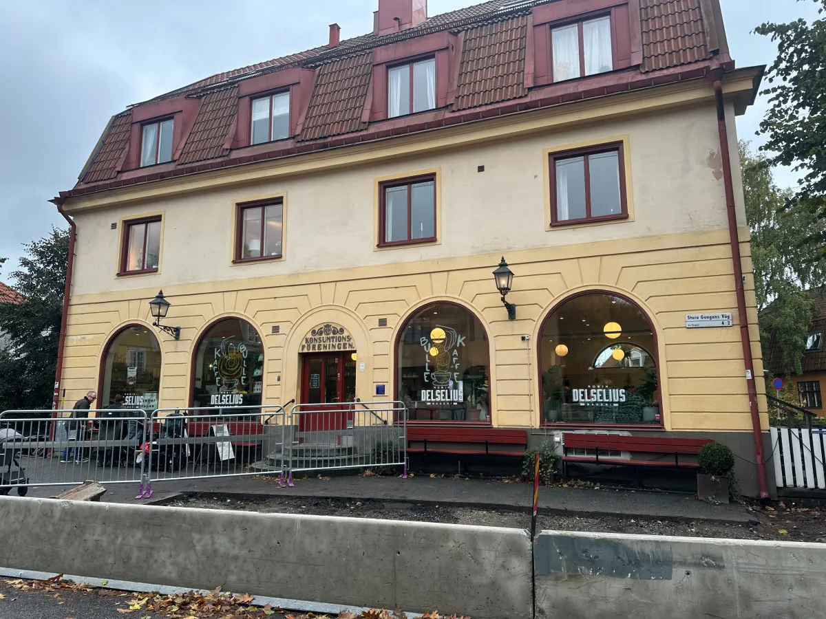 Ett gult hus med rundbågade fönster och skyltar för Delselius. Framför står metallstängsel, och närmast gatan en rad betongblock vid uppgrävd mark.