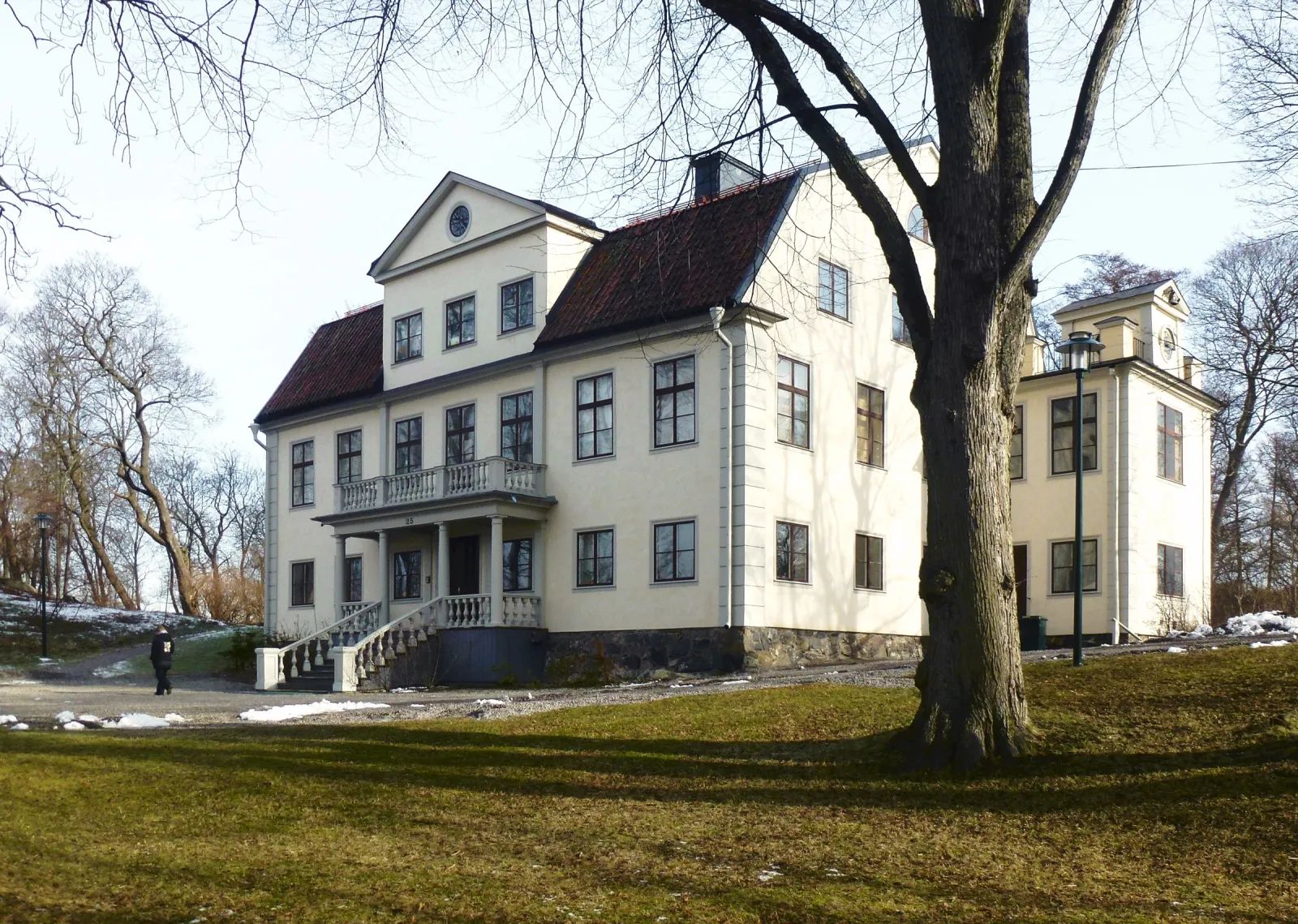 Enskede gårds huvudbyggnad, ett ljust putsat hus i två våningar med fronton och en entréveranda med pelare och balkong. Till höger en tillbyggnad med tornur. Framför står ett stort kalt träd på en gräsmatta med snöfläckar.
